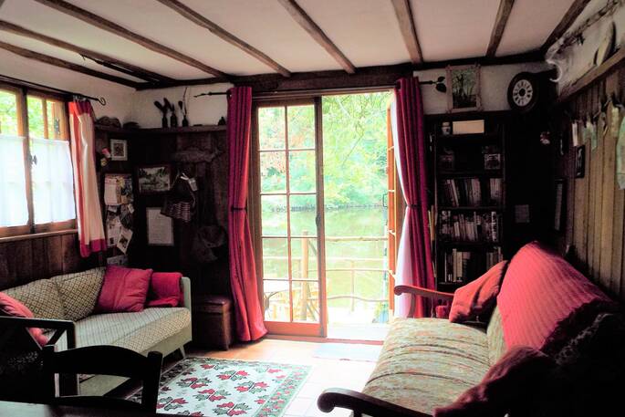 Te cosy living room of the Fisherman's cabin in the Dordogne in France. Cute patterened fabrics cover the comfortable sofas, along with cherry pink pillows and throws. Through the window the gorgeous view on the river and trees can be seen through the window