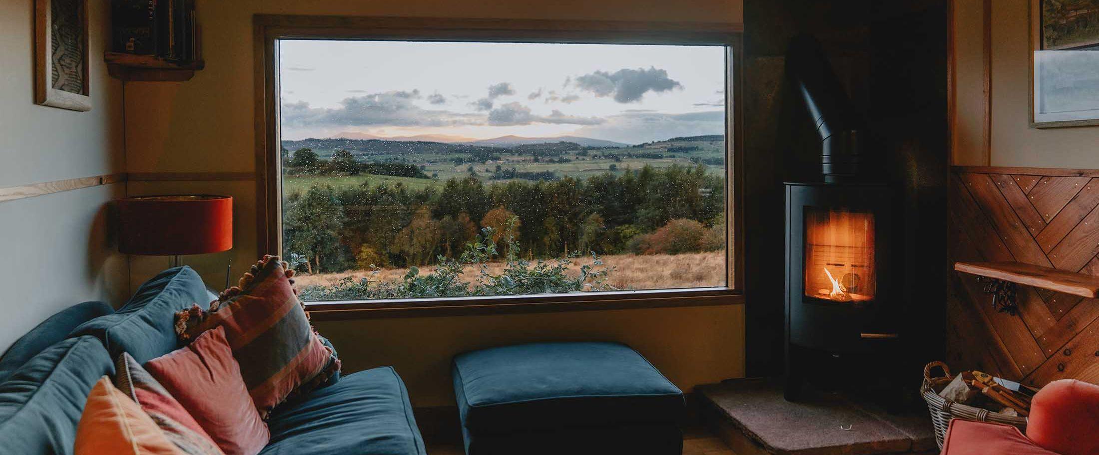 Wood burner in a cabin with a sofa and a window overlooking a forest