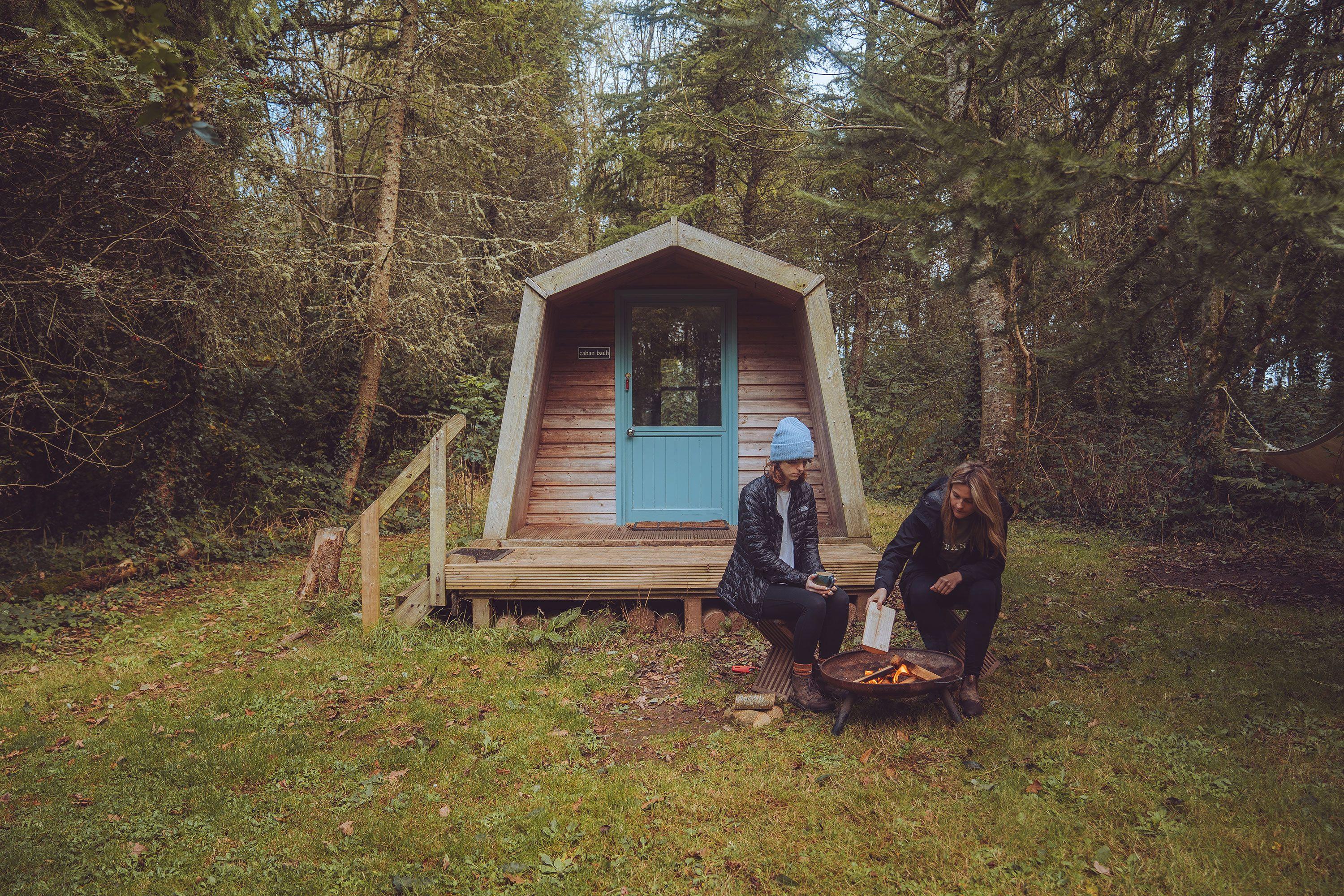 Two people sitting outside a cabin in the woods lighting a fire pit