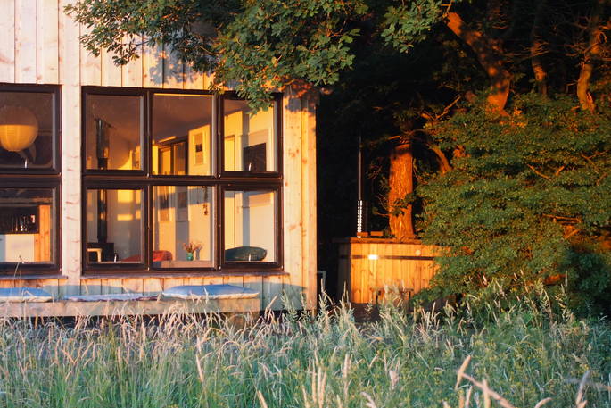 The sun lit cabin  stands in the high grass, pillows lie outside it for lounging or yoga on the decking. Next to the cabin, partially hidden by the tree, is a private wood burning hot tub to soak in after long walks in the coutryside, at Quiet of Stars, Cornwall