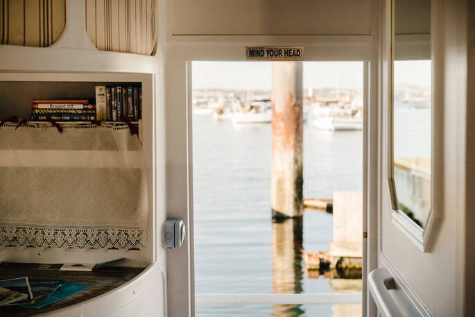 A built-in cabinet holding books and flyers stands next to the door at boat Faithful, through the open door you see the water of Brixham Marina, Devon
