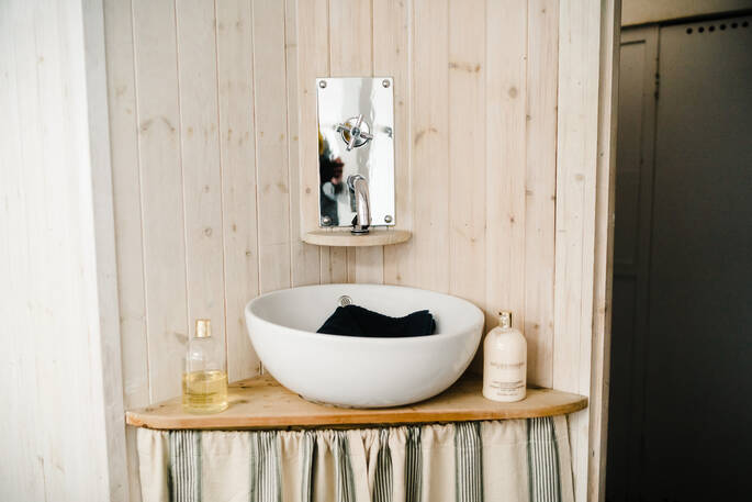 A silver tap is mounted on a wooden wall above a white porcelain sink containing a black towel. Luxurious fragrant soaps are standing alongside it at Faithful, Devon