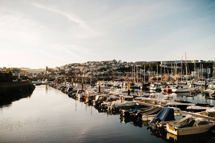 View of the tranquil harbour of Brixham, with lots of little boats moored up on the Brixham Marina. In the background you can see the lovely little town with pastel hued cottages