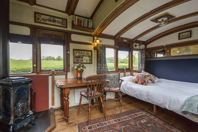 Interior of Camping Coach, Dorset, with vintage decorated cast iron wood burner, antique dining table with chairs, and sofa bed made up as a bed
