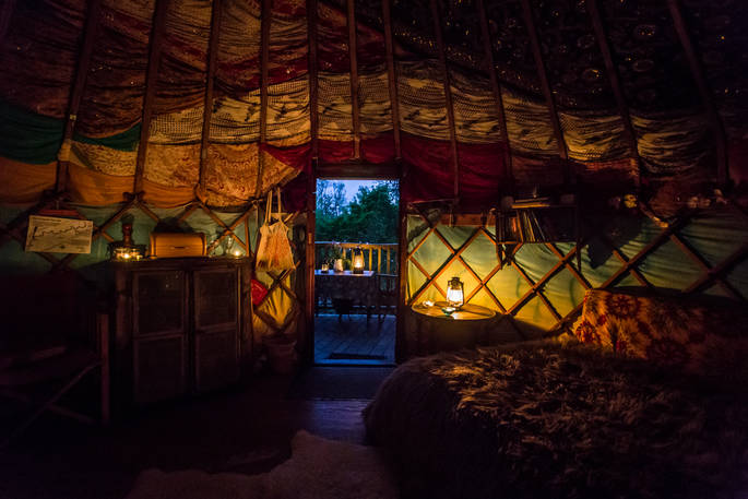 Colourful interior of Old Larch Yurt at dusk illuminated with candles and lanterns and door open leading to raised deck 